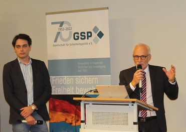 Zwei Männer bei einer GSP-Veranstaltung. Einer spricht am Rednerpult, der andere beobachtet. Ein Banner der Gesellschaft für Sicherheitspolitik.