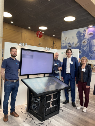 Vier Personen an einem Messestand mit großem Bildschirm und interaktiver Tischplatte, ein Mann hält ein VR-Headset.