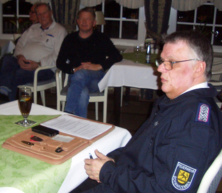 Ein Mann in einer Uniform der 'Feuerwehr Schwarzenbek' sitzt an einem Tisch mit Dokumenten, einem Telefon und einem Getränk, mit zwei anderen Männern im Hintergrund.