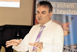 Ein Mann in weißem Hemd und lila gestreifter Krawatte spricht und gestikuliert vor verschwommenem Hintergrund.