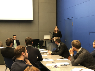 Eine Frau präsentiert auf einem Seminar vor einer Gruppe Männer in einem modernen Konferenzraum mit großer Leinwand.