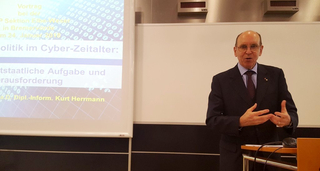 Mann im Anzug, Kurt Herrmann, hält Vortrag über 'Politik im Cyber-Zeitalter' vor einer Leinwand.