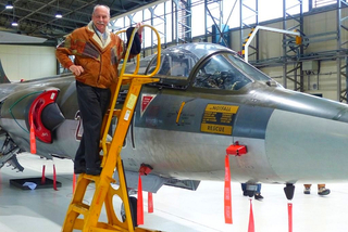 Lächelnder Mann auf gelber Leiter neben grauem Militärjet-Cockpit mit "NOTFALL" und "RESCUE" im Hangar.