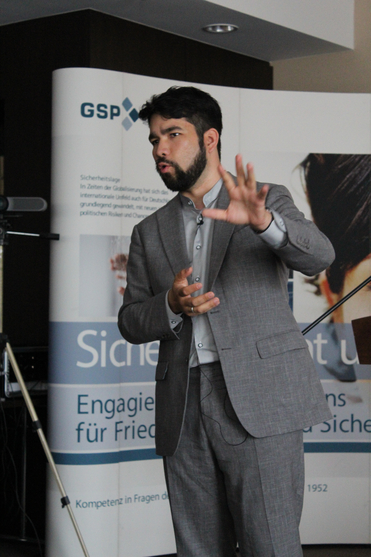 Ein bärtiger Mann im grauen Anzug spricht gestikulierend vor einem weißen Banner mit GSP-Logo und Text.