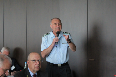 Offizier Heisner spricht ins Mikrofon auf einer Konferenz, gestikulierend. Teilnehmer, einer mit Ukraine-Pin, sind sichtbar.