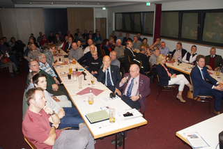 Eine große Gruppe überwiegend männlicher Personen sitzt an langen Tischen in einem Besprechungsraum, viele mit Getränken und Papieren.
