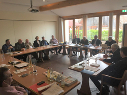 Männer und einige Frauen sitzen an U-förmig angeordneten Holztischen in einem hellen Konferenzraum mit großen Fenstern.