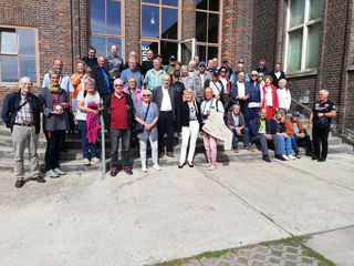 Eine große Gruppe älterer Erwachsener posiert an einem sonnigen Tag vor einem alten Backsteingebäude für ein Foto.