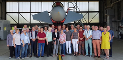 Eine große Gruppe lächelnder Erwachsener posiert für ein Foto vor einem grauen Kampfjet in einem Flugzeughangar.