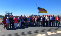 Eine Gruppe Erwachsener auf einem Schiffsdeck mit deutscher Flagge und Marineschiff im Hintergrund unter blauem Himmel.