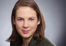 Porträtaufnahme einer lächelnden Frau mit braunen Haaren und blauen Augen, die eine dunkle Jacke trägt und direkt in die Kamera blickt.