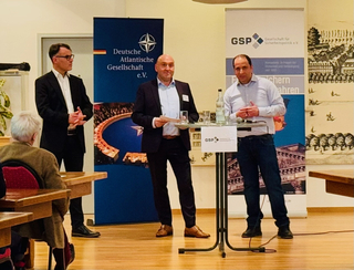 Drei Männer bei einer Diskussionsrunde oder Präsentation vor den Bannern der Deutschen Atlantischen Gesellschaft und GSP.