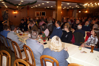 Festliche Versammlung in einem Saal mit vielen Gästen, die einem Redner von Tischen aus zuhören.
