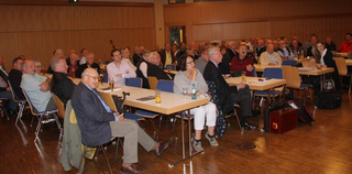 Eine große Gruppe von Erwachsenen und Senioren sitzt an Tischen in einer Halle und nimmt an einer Veranstaltung teil.