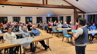 Eine Frau spricht in ein Mikrofon vor einem Publikum älterer Erwachsener, die an Tischen in einem hellen Raum sitzen.