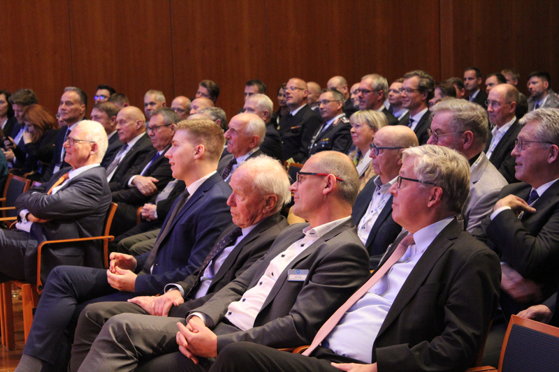 Eine Gruppe von Männern in Anzügen sitzt als Publikum in einem Konferenzsaal und lauscht einer Veranstaltung.