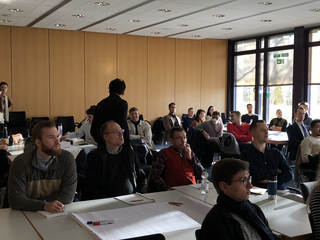 Eine Gruppe von Teilnehmern bei einem Workshop oder Seminar, sitzend an Tischen in einem hellen Raum mit Fenstern und Holzvertäfelung.