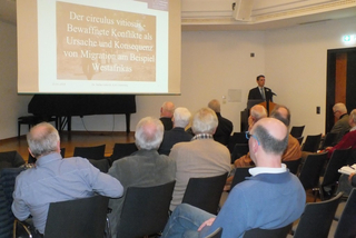 Redner an einem Pult vor Publikum in einem Hörsaal. Ein Bildschirm zeigt eine Präsentation über bewaffnete Konflikte und Migration in Westafrika.