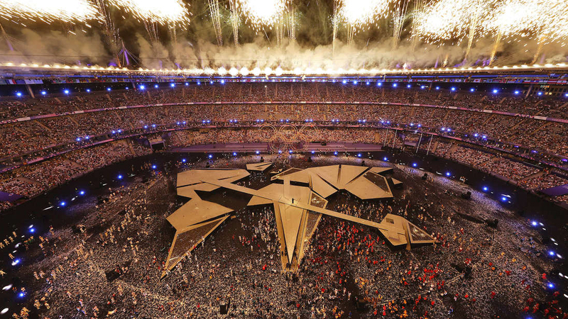 Feuerwerk über einem gefüllten Olympiastadion mit leuchtenden Ringen, geometrischer Bühne und Darstellern auf dem Feld.