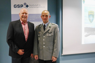 Ein Mann im Anzug und ein Soldat in Uniform vor einem GSP-Banner und einer NATO-Präsentation.