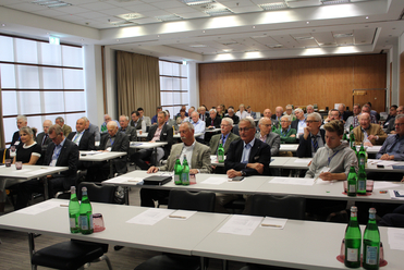 Eine große Gruppe von Personen, hauptsächlich Männer, sitzt an Tischen mit Wasserflaschen und hört bei einer Konferenz zu.