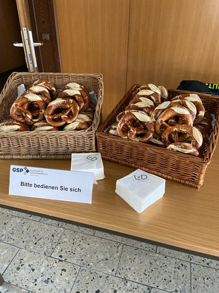 Zwei Weidenkörbe voller gesalzener Brezeln, Serviettenstapel und ein GSP-Schild mit der Aufschrift „Bitte bedienen Sie sich“ bei einer Veranstaltung.