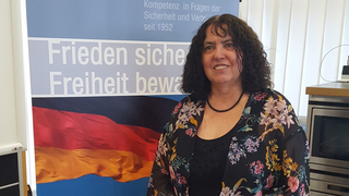Lächelnde Frau in geblümter Bluse vor einem Banner mit der deutschen Flagge und „Frieden sichern, Freiheit bewahren“ Text.
