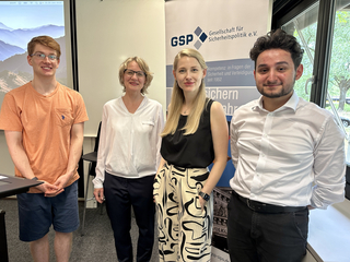 Vier lächelnde Personen, zwei Männer und zwei Frauen, stehen vor einem Banner der Gesellschaft für Sicherheitspolitik (GSP).