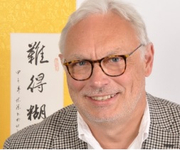 Ein lächelnder, grauhaariger Mann mit Brille in einer gemusterten Jacke vor gelbem Hintergrund mit Kalligraphie.