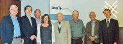 Eine Gruppe von sieben Männern und einer Frau posiert für ein Foto bei einer Veranstaltung, mit einem GSP-Banner im Hintergrund.