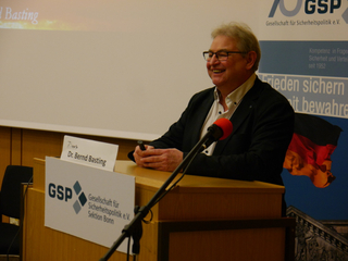 Dr. Bernd Basting lächelt an einem Rednerpult mit Mikrofon, GSP-Banner und deutscher Flagge im Hintergrund.