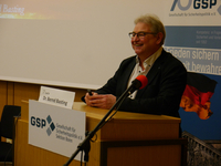 Dr. Bernd Basting lächelt an einem Rednerpult mit Mikrofon, GSP-Banner und deutscher Flagge im Hintergrund.