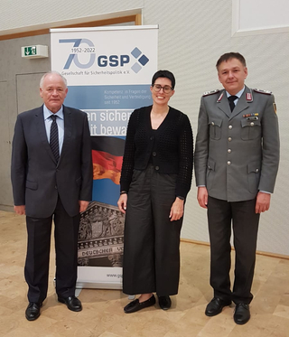 Drei Personen, darunter ein Offizier, stehen vor einem Banner der Gesellschaft für Sicherheitspolitik (GSP).