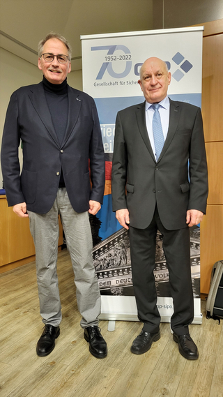 Zwei Männer, einer in dunklem Blazer und grauer Hose, der andere im dunklen Anzug mit blauer Krawatte, stehen vor einem Banner.