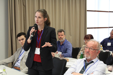 Junge Frau hält ein Mikrofon und spricht vor Publikum bei einer Konferenz, andere Teilnehmer sitzen und hören zu.