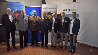 Sieben Männer, teils in Uniform, Vertreter deutscher Verteidigungs- und Sicherheitsorganisationen, posieren vor Bannern.