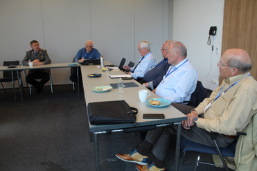 Sechs Männer, einer in Militäruniform, sitzen an einem Konferenztisch mit Snacks und Getränken in einem modernen Raum.