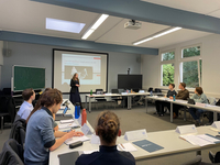 Eine Rednerin präsentiert Studenten in einem hellen Klassenzimmer zum UN-Sicherheitsrat, mit Projektor und Tafel.