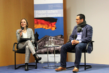 Zwei Redner auf einer Bühne während einer Diskussion, mit einem Banner der deutschen Flagge und des Reichstagsgebäudes im Hintergrund.