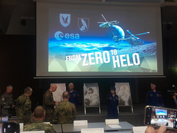 Ein Bildschirm zeigt 'FROM ZERO TO HELO', ESA-Logo, Helikopter und eine Mondlandschaft. Militärpersonal und Astronauten bei einem Event.