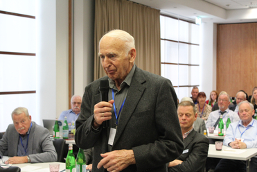 Ein älterer Mann mit weißen Haaren spricht bei einer Konferenz ins Mikrofon, im Hintergrund sitzt das Publikum an Tischen.
