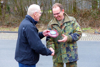 Zwei lächelnde Männer, einer zivil, der andere in Militäruniform, schütteln Hände bei einer Auszeichnungsübergabe.