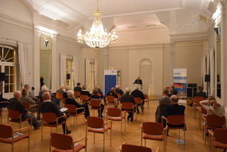 Ein Redner spricht vor Publikum in einem großen Saal. Veranstaltung mit Bannern von GSP und Konrad-Adenauer-Stiftung.