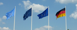 Vier wehende Flaggen: Vereinte Nationen, NATO, Europäische Union und Deutschland, vor blauem Himmel mit Wolken.