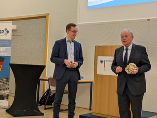 Zwei Männer präsentieren, der ältere hält ein helles, strukturiertes Objekt, der jüngere hört zu. Im Hintergrund der Bundesadler.
