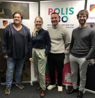 Vier lächelnde Personen, drei Männer und eine Frau, vor Bannern des Think Tanks Polis 180 und militärischen Symbolen.