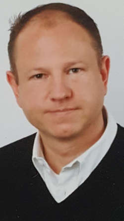 Kopfaufnahme eines Mannes mit kurzen dunklen Haaren, der ein weißes Hemd und einen schwarzen V-Pullover trägt.