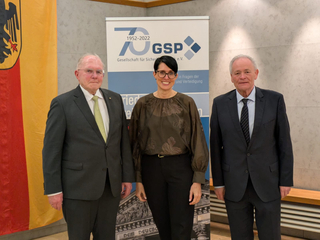 Drei Personen, eine Frau in der Mitte und zwei Männer, stehen lächelnd vor einem GSP 70-Jahre-Jubiläumsbanner.