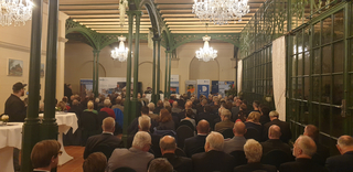 Zuschauer bei einer Konferenz in einem festlichen Saal mit Kronleuchtern und Säulen, lauschen einem Redner im Hintergrund.