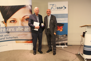 Zwei Männer in Anzügen posieren vor GSP-Bannern, einer hält eine Urkunde und eine Medaille.
